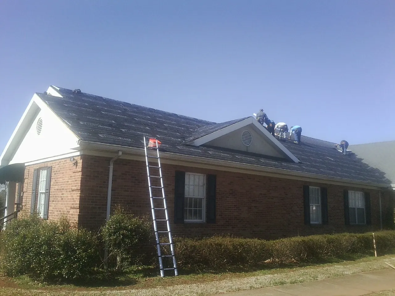 Crew tearing shingles off a brick ranch roof with a ladder set — Emergency Roof Repair work by our crew in Sanford